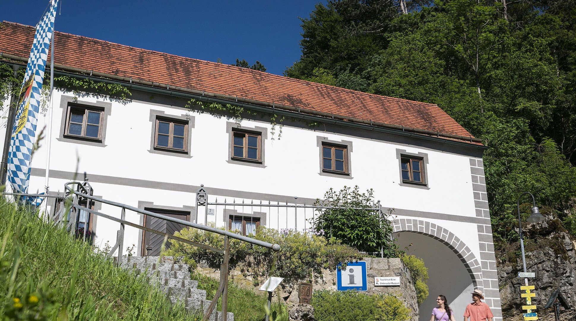 Weißes Gebäude mit rotem Ziegeldach, bayerischer Flagge und zwei Personen vor einem Torbogen bei Sonnenschein