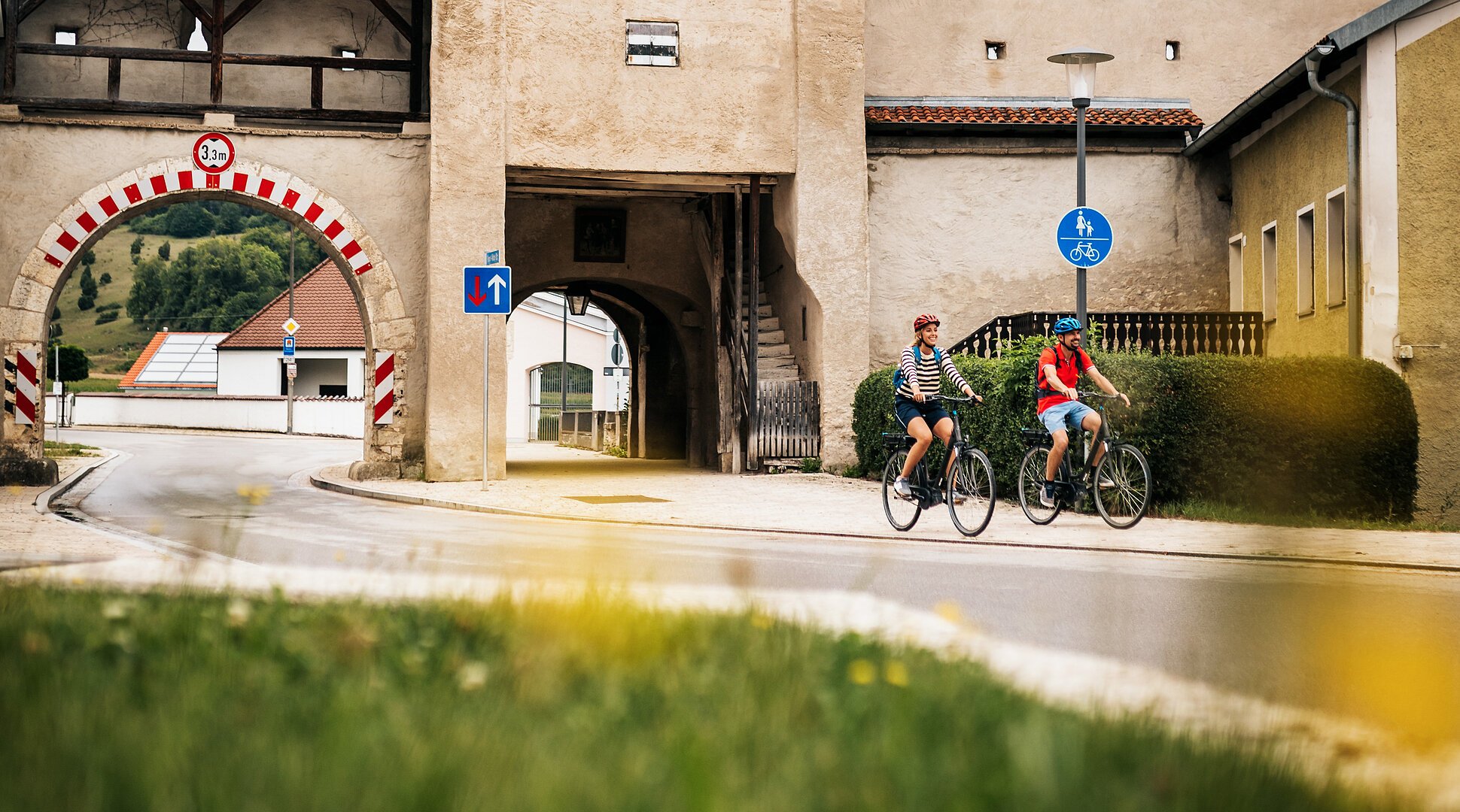 Zwei Radfahrer fahren an einem historischen Stadttor mit rotem Dach und Durchfahrt vorbei.
