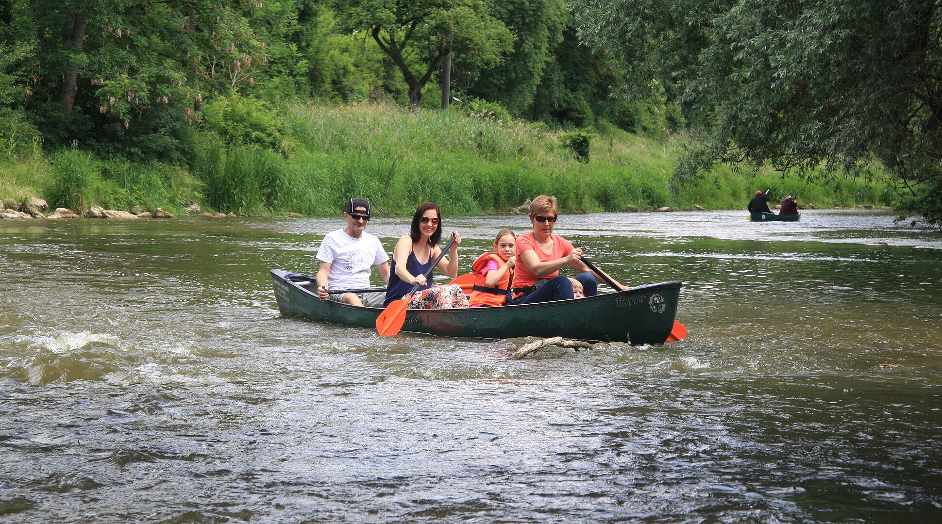 Eine Familie ist einem Kanu vom Schmidt Bootsverleih auf der Altmühl unterwegs. Das Kanu steht waagrecht im Wasser und alle schauen uns an. Im Hintergrund ist das grüne Ufer und Bäume zu sehen.