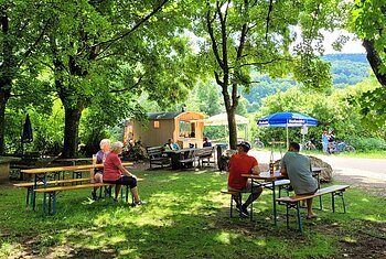 Mein Platz an der Bruck Menschen sitzen an Holzbänken unter Bäumen in einem schattigen Biergarten mit Imbisswagen im Hintergrund.